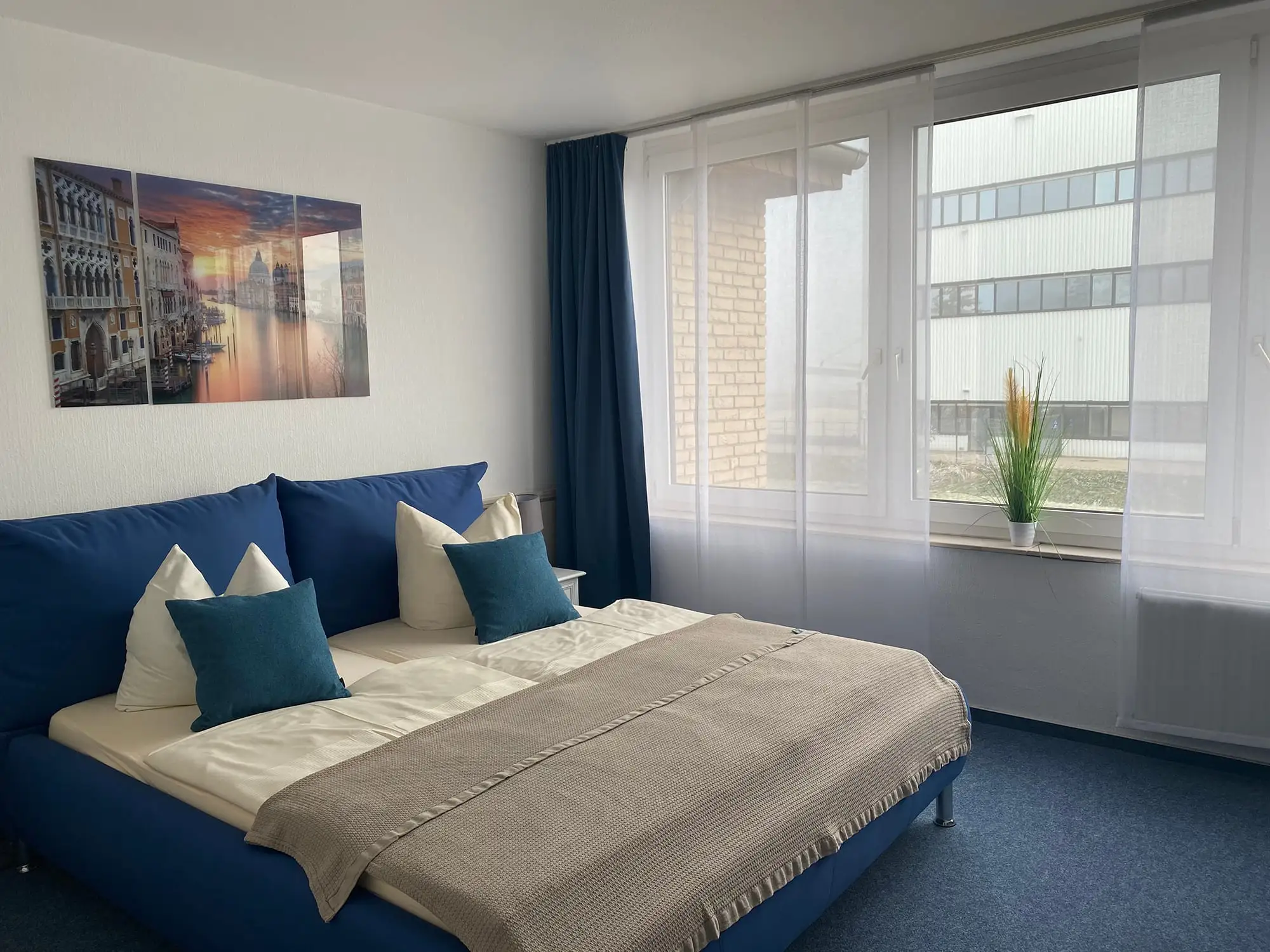 Hotelzimmer mit Doppelbett, blauem Kopfteil, petrolfarbenen Kissen und Bild von Venedig