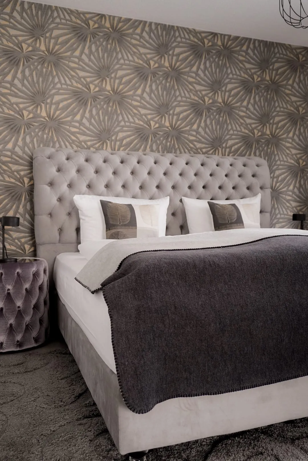 Kingsize-Bett mit hohem Chesterfield-Kopfteil vor grafischer Ornamenttapete, daneben ein gepolsterter Nachttisch